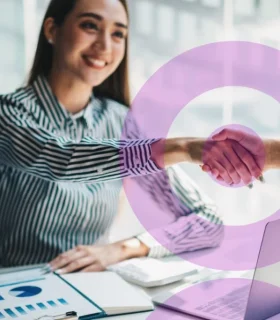Mulher sorridente cumprimentando outra pessoa com um aperto de mão em ambiente corporativo. Sobre a imagem há elementos gráficos em rosa com formato circular. Na mesa, estão um notebook, relatórios com gráficos e uma caneta, sugerindo uma reunião de negócios ou fechamento de parceria.