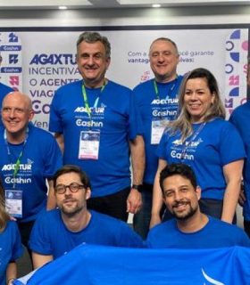 Integrantes dos times Cashin e Agaxtur posam sorridentes para foto em estande de evento, vestindo camisetas azuis com as respectivas marcas. Ao fundo, banners destacam informações sobre prêmios, incentivos digitais e oportunidades de negócios.