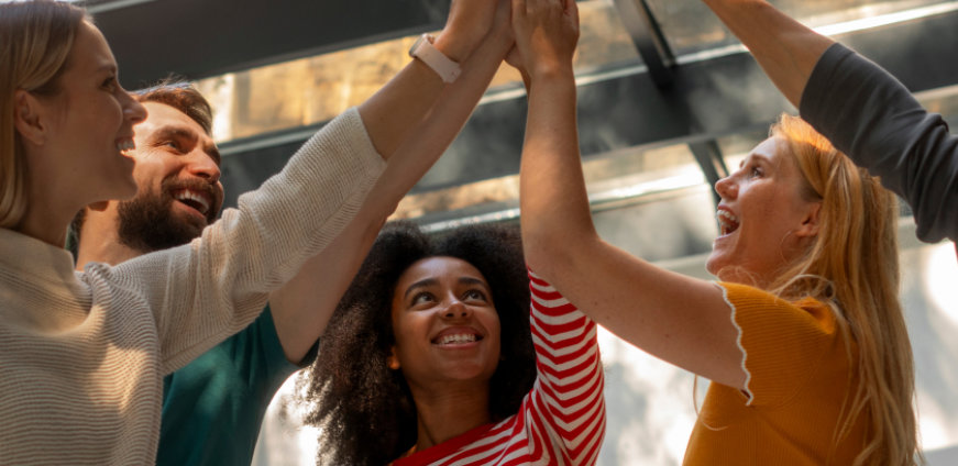 Grupo de cinco pessoas sorridentes comemorando com um high five coletivo em um ambiente iluminado, simbolizando união, trabalho em equipe e celebração de conquistas