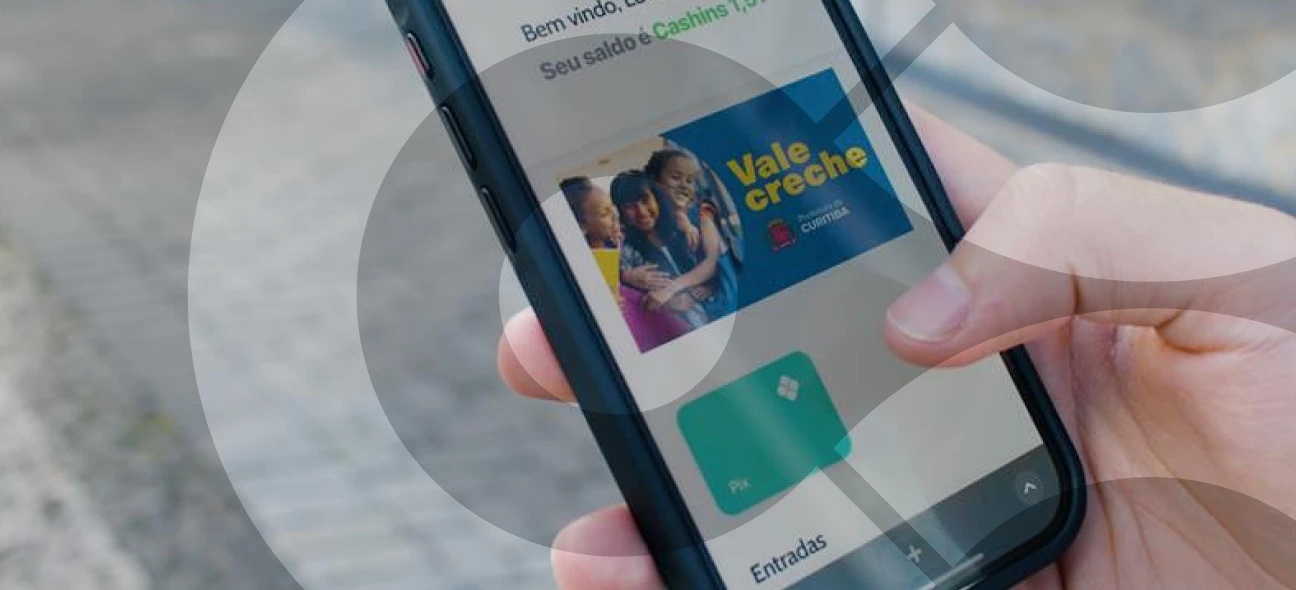 Imagem de uma mão segurando um smartphone que exibe a tela do aplicativo Cashin, mostrando o saldo disponível e a opção "Vale-Creche" da Prefeitura de Curitiba. 